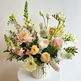 A floral arrangement featuring a variety of flowers in shades of white, peach, and green, placed in a white ceramic pot.