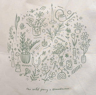 A custom tote bag featuring a circular design with various illustrated plants and a pot, with the text 'The Wild Pansy' and the artist's signature, LeeAndra Cianci.
